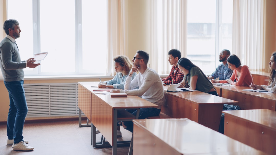 Male professor in casual clothes is talking to group of students sitting at tables in classroom and making notes. Large lectu
