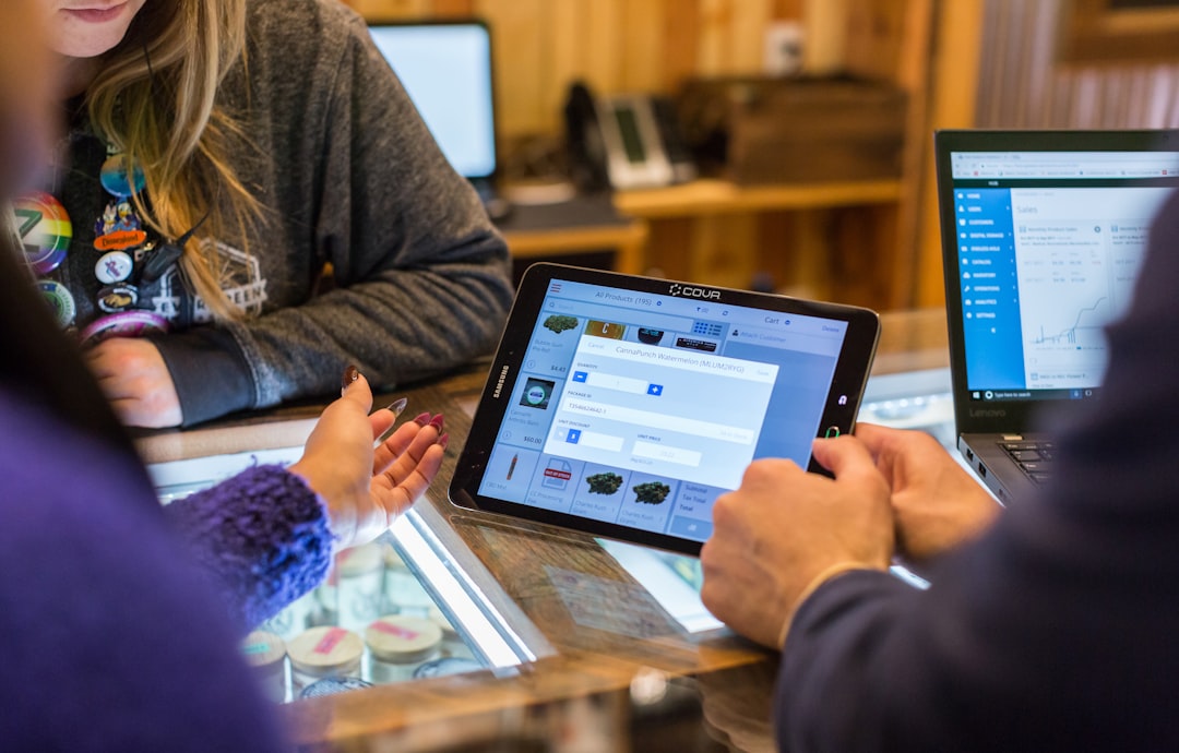 Two cannabis customers discuss product options with a female budtender across a dispensary sales counter. Between them is a l