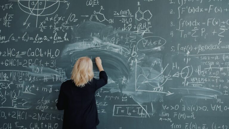 Adult woman teacher in elegant suit and glasses is writing formulas on blackboard talking teaching class at school. Education