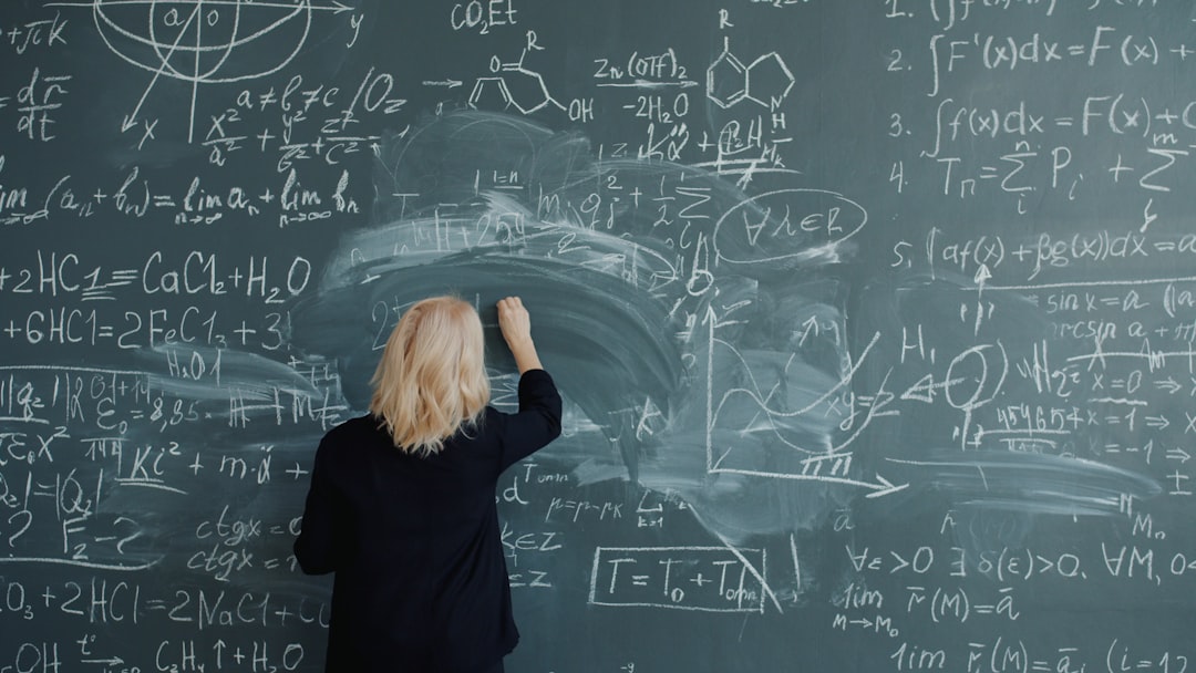 Adult woman teacher in elegant suit and glasses is writing formulas on blackboard talking teaching class at school. Education