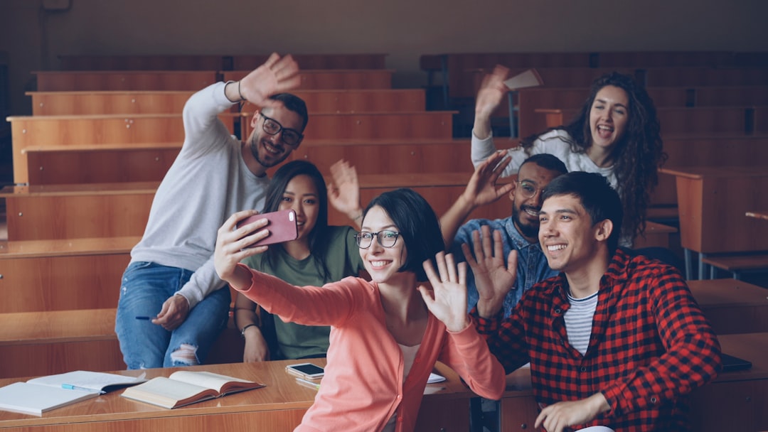 Joyful students are having fun recording funny video in classroom waving hands and smiling sitting at desks in lecture hall. 