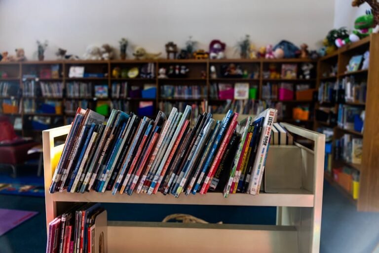 A warm library interior featuring a book cart in focus with colorful children's books on shelves.