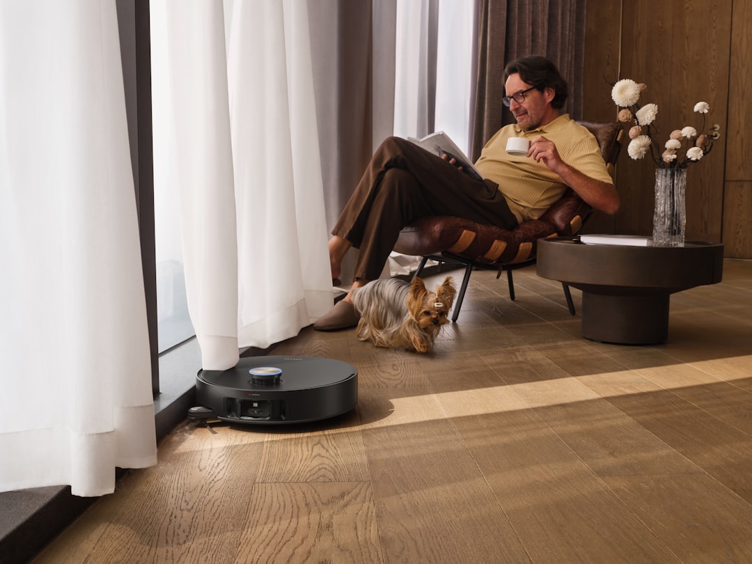A man relaxes comfortably in a stylish armchair by a sunlit window, reading a book and enjoying a warm drink, while a robot v