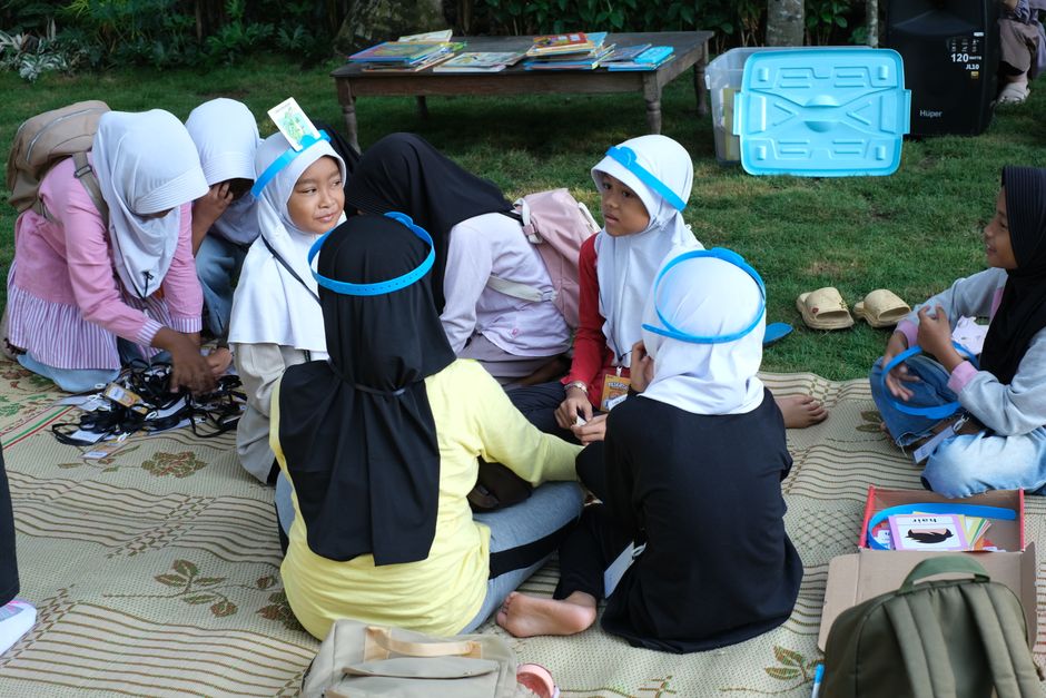 A group of children sitting on a mat outdoors engaged in a group activity wearing headbands.