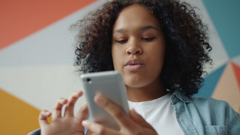 Attractive young girl is holding smartphone touching screen relaxing at home on colorful background. Modern technology, inter