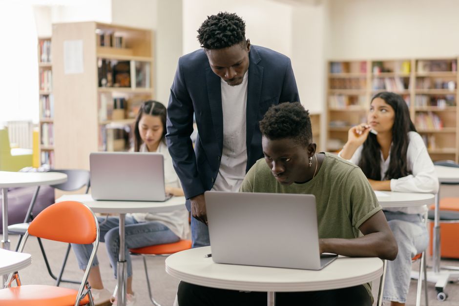 Students learning in a classroom setting with a teacher assisting and laptops on desks, creating an interactive education env