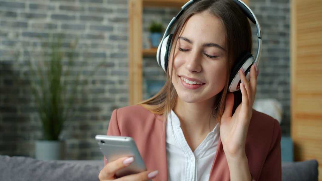 Happy student attractive girl is listening to music in headphones holding smartphone touching screen sitting on sofa at home.