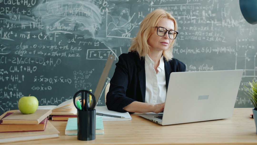 Happy teacher attractive matude adult is smiling using laptop in class typing working with chalkboard in background. People a