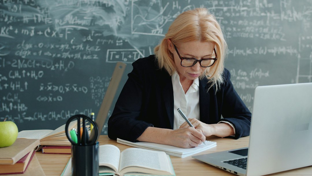 Mature woman in suit working in class writing sitting at desk with books and laptop, blackboard with formulas is visible in b