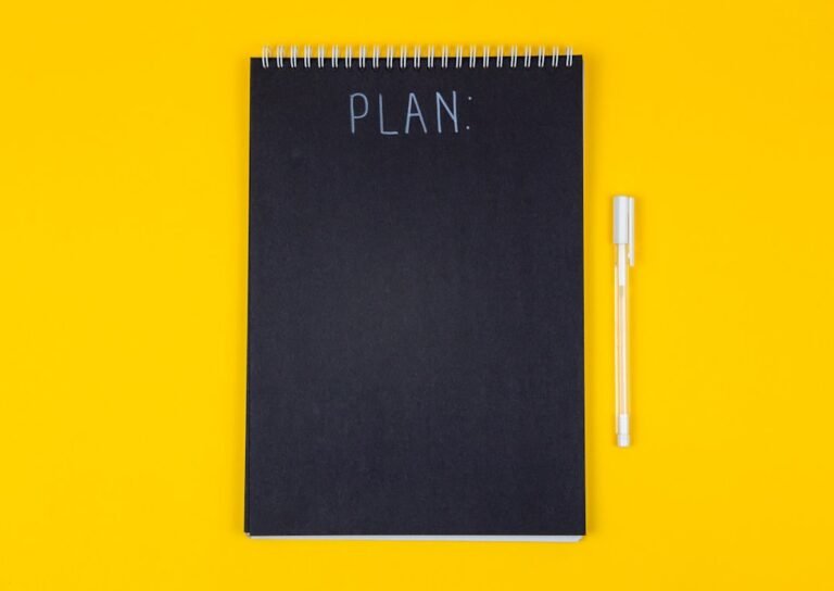 Wide view image of blank black spiral note pad and white marker with calligraphic inscription plan on yellow background