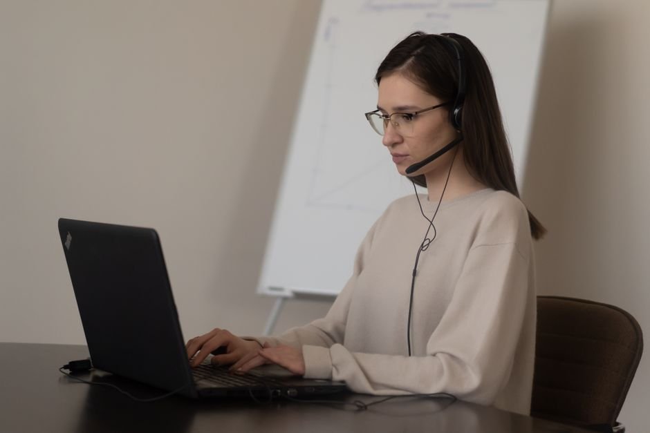 Serious young woman in casual outfit with eyeglasses sitting on chair at table and working on laptop with headphones with mic