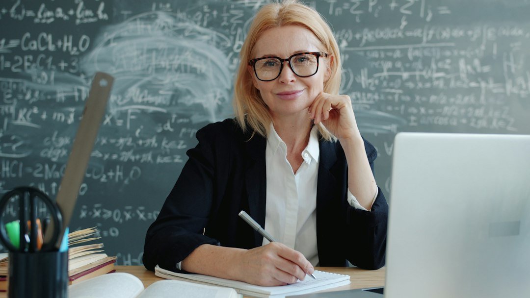 Friendly mature lady is smiling sitting at desk in classroom looking at camera indoors, chalkboard with formulas is visible i