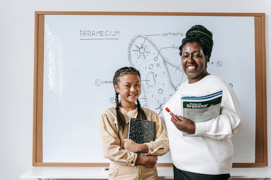 Teacher and student holding a grammar workbook in front of a classroom whiteboard