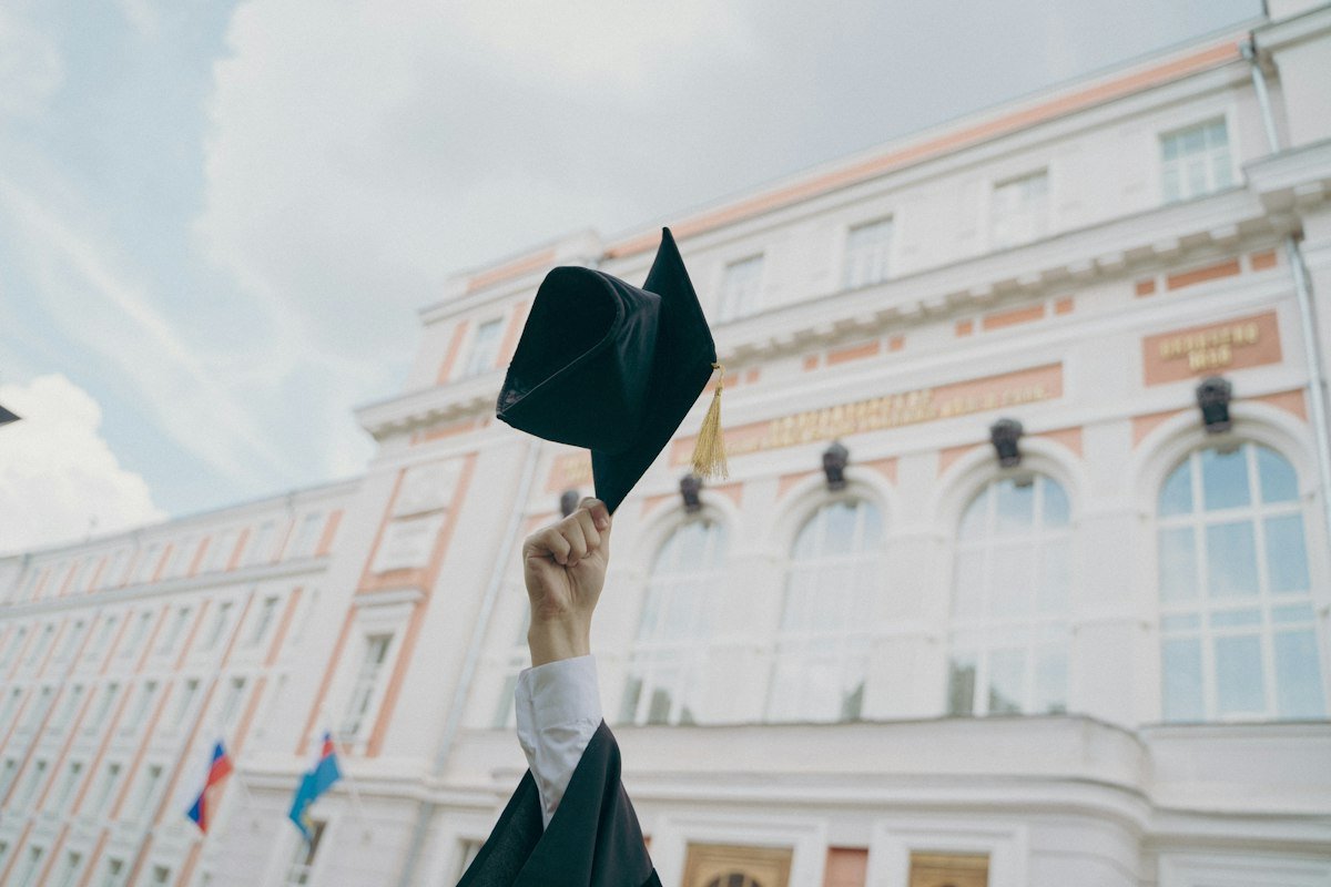 A hand holding a graduation cap in front of a university building, illustrating quick study and school of hard knocks